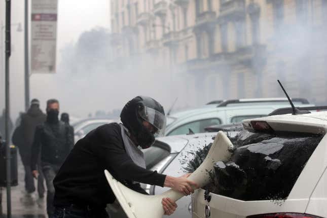 A rioter smashes an object into the rear window of a car in Milan.