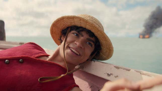 A One Piece live action still shows Luffy smiling while a ship burns in the background. 