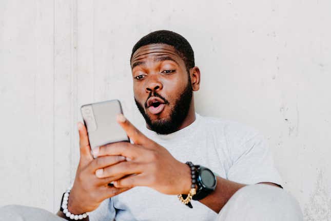 Young african american man with surprised facial expression hold smartphone in his hands, typing message, chatting with friends in social networks, playing online game, isolated against grey wall