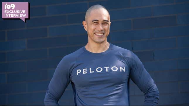 Peleton instructor/host Sam Yo poses in a longsleeve, blue Peleton shirt in front of a blue brick wall.