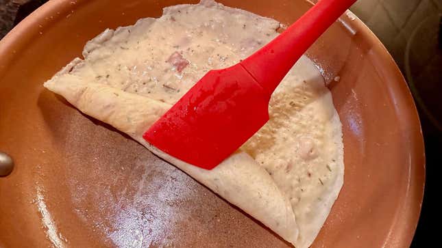 A red rubber spatula is rolling a thin pancake in a frying pan.