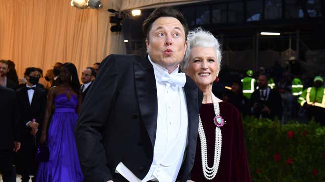 Elon Musk in a tuxedo makes a kissy face to the side standing next to his mother.