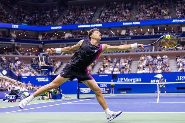 American Ben Shelton, wearing black shorts and a black top with bright pink accents, stretches his racquet to return a ball at center court.