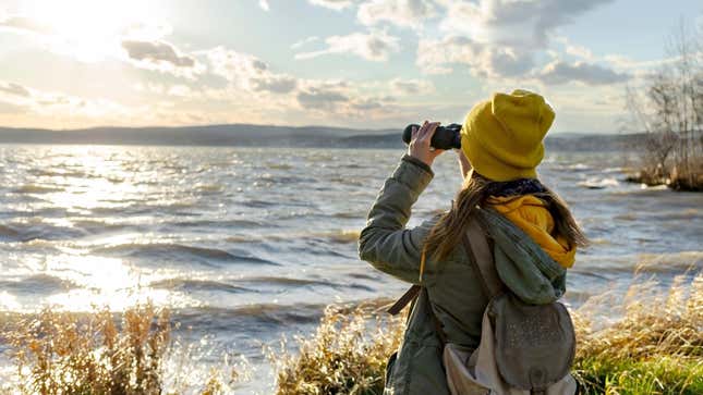 A woman in hiking gear  looks out across a lake using binoculars