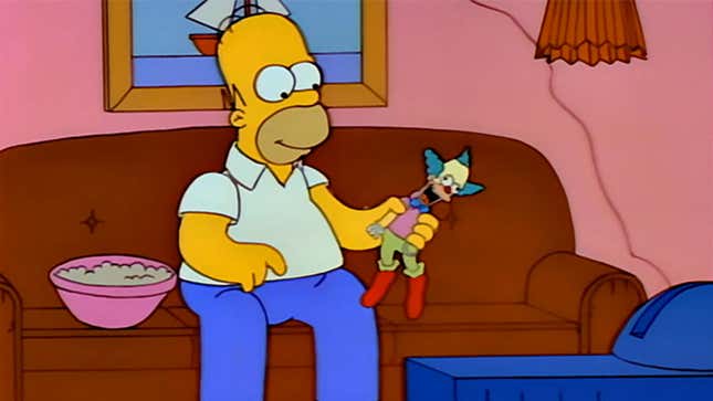 A screenshot from The Simpsons shows Homer holding a comedian doll. 