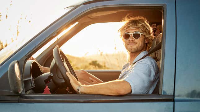 Porträt eines selbstbewussten Mannes mittleren Alters, der bei Sonnenuntergang einen Lieferwagen fährt.  Lächelnder Mann trägt während der Autofahrt eine Sonnenbrille.  Er hat blondes Haar.