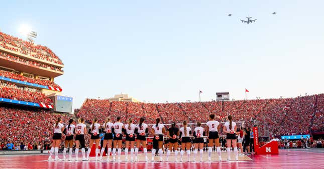 A KC-135 and three F-16 jets fly over a packed Memorial Stadium in Nebraska, where the women's volleyball team stands in a line with their backs to the photographer.