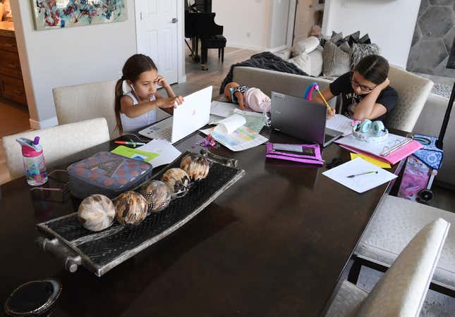 Two young girls sit at a dining room table working on their computers for remote learning during a lockdown.