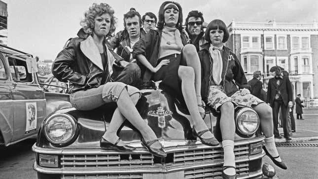 A black and white photo of young people sat on a car hood. 