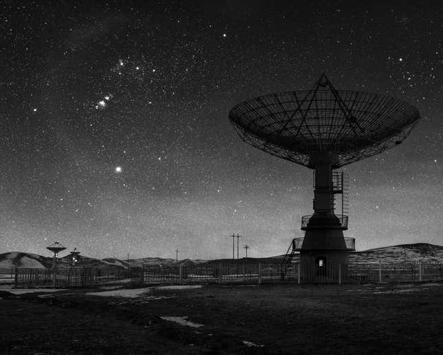 The silhouette of a radio telescope, the cosmos behind it, in black and white.