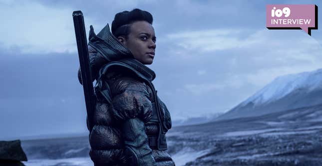 A woman (Leah Harvey's Salvor Hardin) wearing a parka with a weapon strapped to her back stands against a desolate backdrop in a scene from Foundation.
