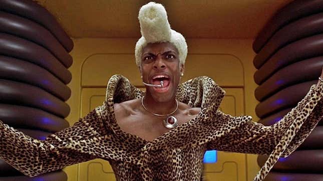 Ruby Rhod, in a giant yellow pompadour and leopard print onesie, screams into a microphone.