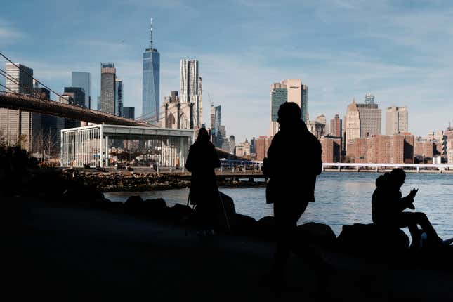 Figures are in silhouette on the waterfront with a view of the Brooklyn Bridge and Manhattan skyline in the distance.