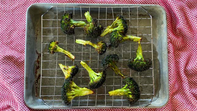 Stalks of roasted broccoli rest on a wire rack suspended over a shallow baking pan placed on a red checked cloth