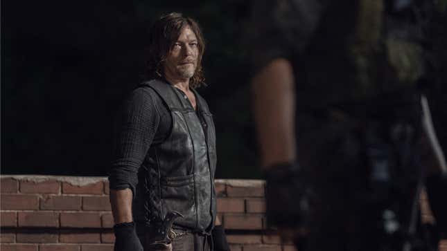 Daryl, in a black leather vest and long-sleeved shirt, stands in front of a low brick wall at night.