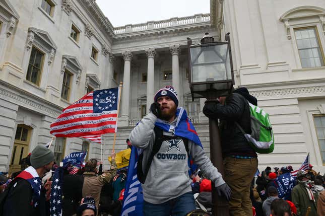 A man wearing a trump flag as a cape holds a phone to his ear during the Jan. 6 insurrection.