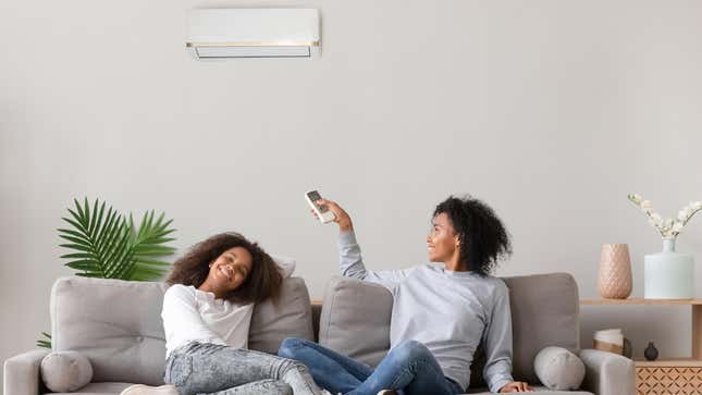 Photograph of a Black teenage girl and Black woman relaxing on a gray sofa. The woman points a remote control at a wall-mounted air conditioning unit, and there's a green palm plant behind the couch.