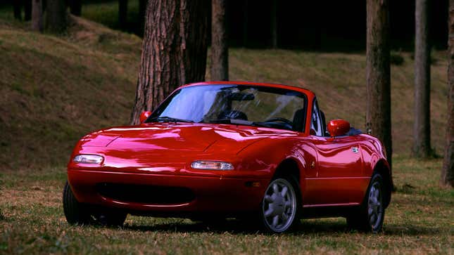 A photo of a red Miata parked in a forrest. 