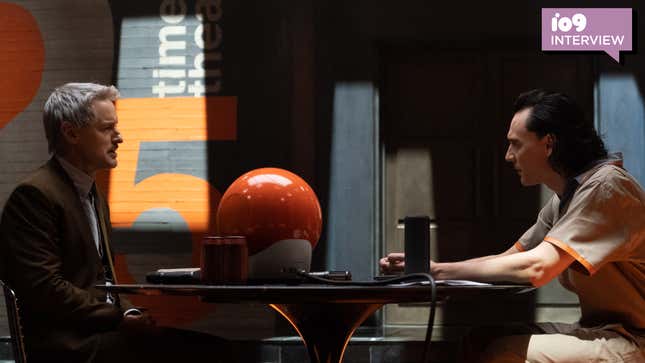Owen Wilson's Agent Mobius in his TVA suit, sits across a table from Tom Hiddleston's Loki in his prison garb with a sphere-like TV in between them.