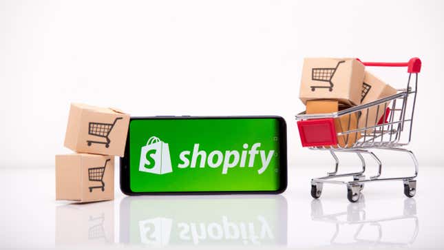 A phone with the shopify logo in front of a few boxes and a shopping cart.