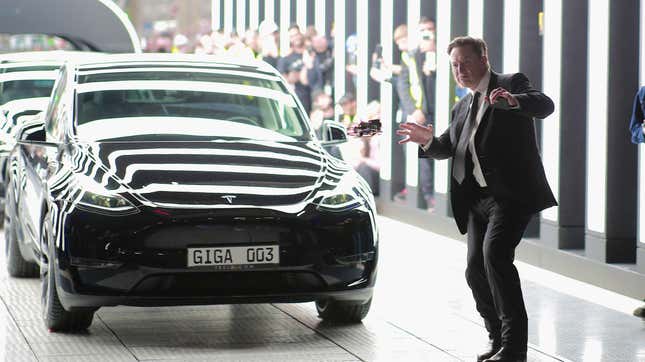 A photo of Elon Musk in a suit making weird hand gestures in front of a black Tesla.
