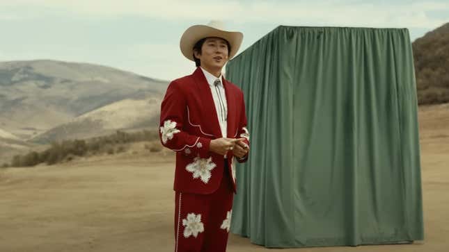 Steven Yeun in a cowboy outfit standing in front of a curtain-covered box.
