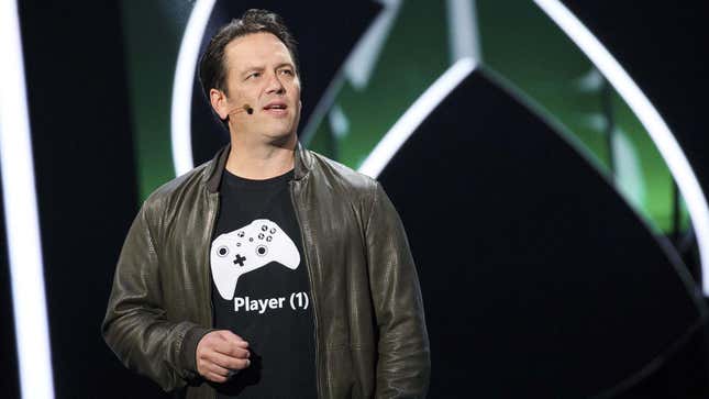 Phil Spencer stands on stage in front of a large black and white Xbox logo. 