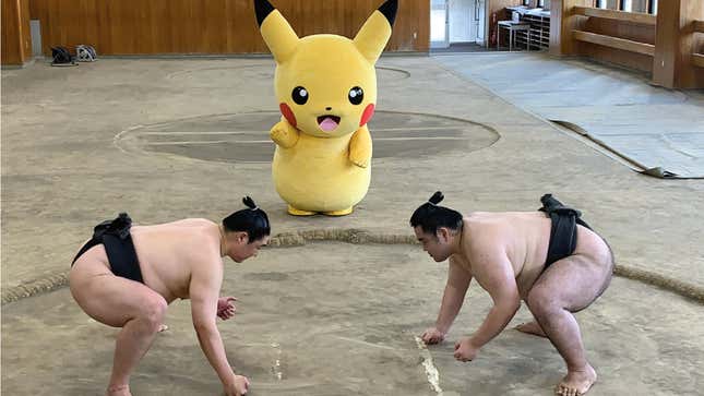 Two sumo wrestlers face off in front of a person wearing a full-body Pikachu costume. 