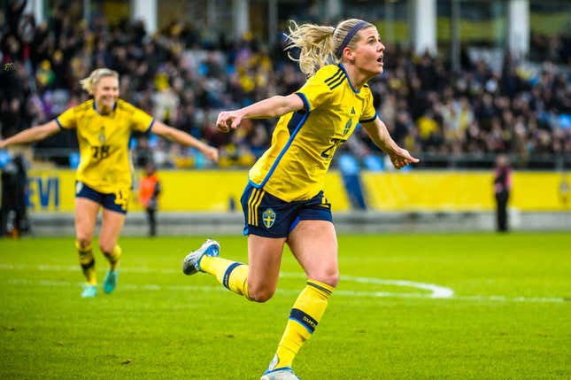 A white woman with a blonde ponytail in a yellow jersey, blue shorts, and yellow socks runs across a soccer pitch with her arms out wide.