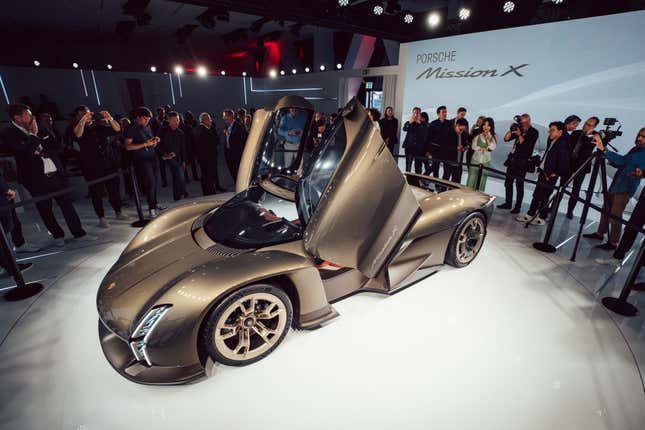 Overhead shot of the Porsche Mission X. People gather behind a velvet rope to take pictures. on a white mirrored stage surrounded my stage lights in the rafters. A huge digital display behind the silver-gold car reads Porsche Mission X