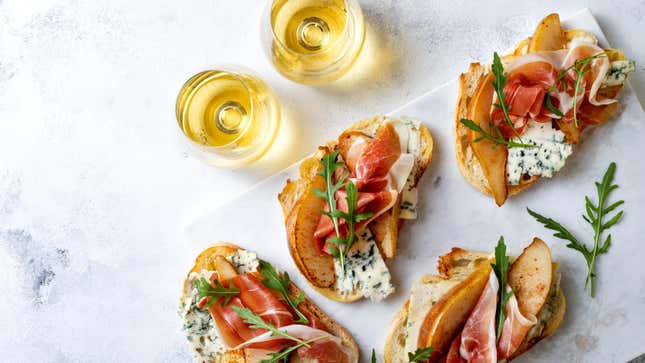 Four prosciutto, pear, and blue cheese crostini sit on a white slate next to two glasses of sparkling wine on a white table.