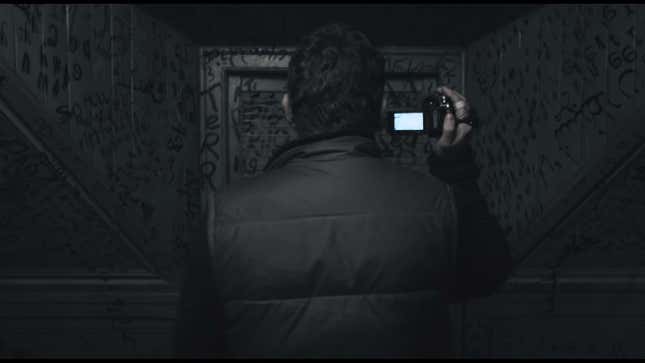 In a screenshot from Book of Blood, a man holds a camcorder in a dark hallway with walls covered in strange writing