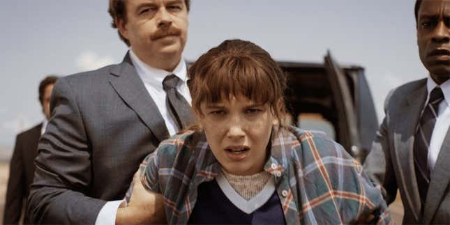 Millie Bobby Brown's Eleven, looking quite a bit older and wearing a flannel shirt, is being held back by the arms by two men in grey suits.