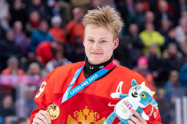 A smiling young white man with spiky blonde hair, wearing a red hockey jersey, shows off the gold medal around his neck.