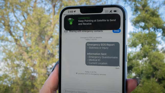 An iPhone 14 in front of trees showing Emergency Satellite connectivity with Apple's SOS system.