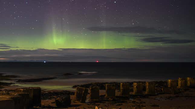 A beach with a row of standing stones looking out over the water with the Aurora Borealis above.