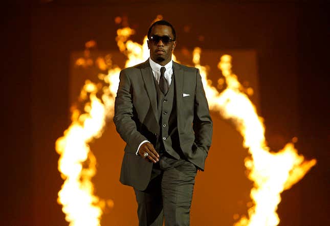 Sean P. Diddy Combs walks a runway in a dark suit, with fire blazing in the background. 