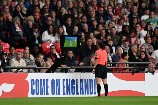 A referee in a neon red shirt stands with her back to the camera, watching the VAR monitor. The audience is waiting expectantly in the stands in front of her. The wall of the football pitch is emblazoned with the message: "Come on England!"