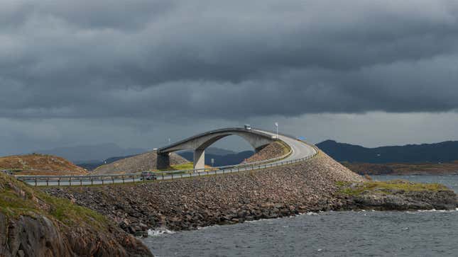 A photo of a swooping road bridge in Scandinavia with dark clouds and mountains behind. 