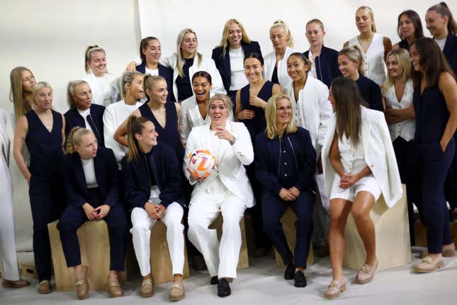 A group of women, all wearing white and black, smile and laugh while the women at their center holds a soccer ball. 