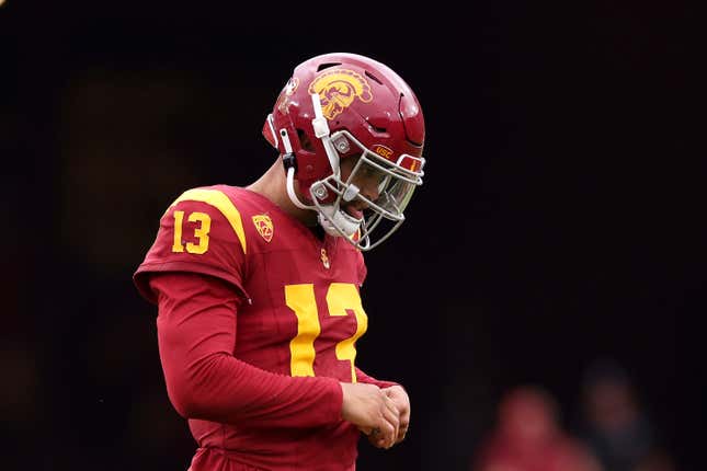 USC quarterback Caleb Williams, dressed in a red uniform with yellow numbers, folds his hands while looking down at something on a football field.