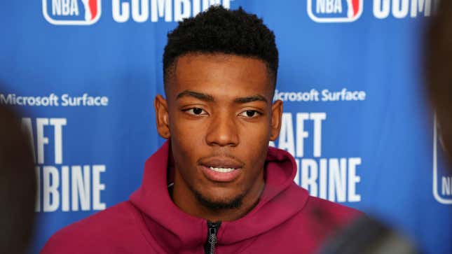 A young Black man in a red hoodie is pictured in talking to reporters in front of a blue background at the NBA draft combine.