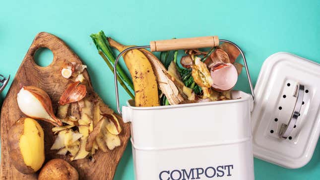 Top-down photograph of food waste on a robin's egg blue background. A wooden cutting board with partially peeled potatoes and onions lies next to a white rectangular bucket labeled "COMPOST." The compost bucket contains banana peels, egg shells, kale stems, and other food scraps. 