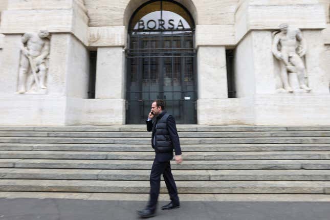 A man dressed in black talks on the phone while passing in front of the white stone steps of the Italian Stock Exchange in Milan.
