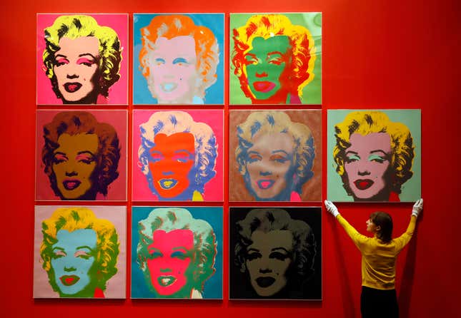 10 Andy Warhol screen prints of Marily Monroe being hung on a red wall by a woman.