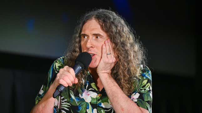 An image of Weird Al Yankovic with a microphone to his month and holding his face.