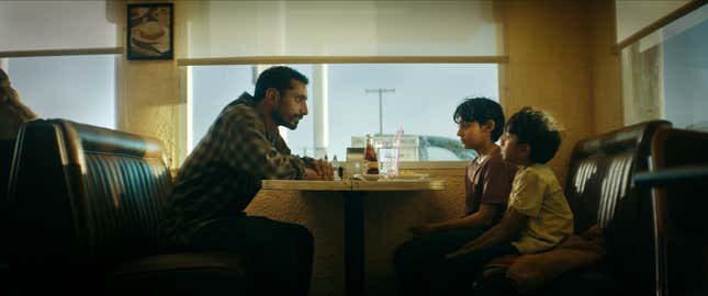 A father and his two young sons sit across a table in a diner, looking very intensely at each other.