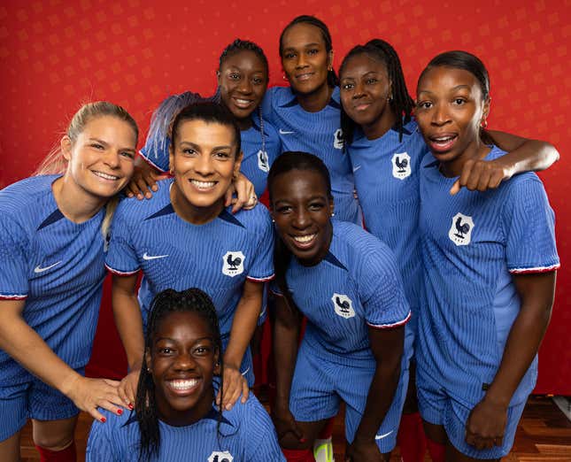 A group of women in matching blue shirts and shorts smile for the camera as part of a team photo in front of a red background.