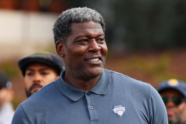 A middle-aged Black man with salt and pepper curly hair and a blue polo shirt with a Knicks logo in the chest smiles.