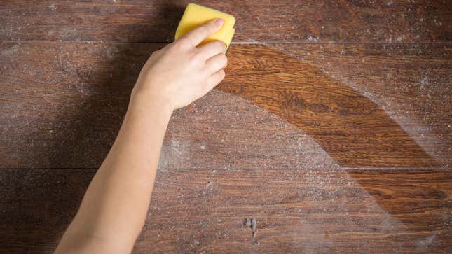 A person wipes dust off a wooden table with a yellow sponge